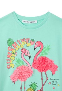 Maglietta verde menta con una stampa colorata di due fenicotteri rosa con dettagli in pompon e testo che recita "SUNSHINE" in un font giocoso.