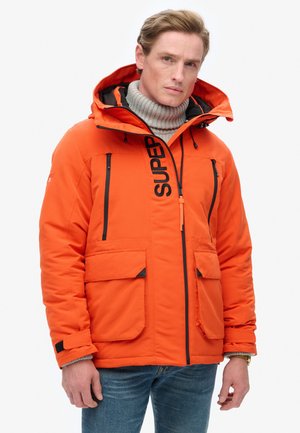 Homme blond portant un manteau d'hiver à capuche orange vif avec des fermetures éclair noires, de grandes poches avant, et un pull à col roulé gris en dessous.