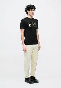 Fiatal férfi göndör hajjal, fekete EA7 Emporio Armani pólóban, krémszínű szöveges melegítőnadrágban és fekete sportcipőben, fehér háttér előtt állva.