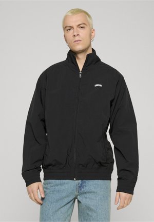 Ung person med kort blondt hår iført en svart glidelåsjakke med "UNFAIR"-logo og lyseblå jeans, stående mot en ensfarget bakgrunn.