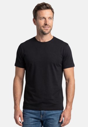 Man met kort bruin haar, gekleed in een effen zwart T-shirt met korte mouwen en een blauwe spijkerbroek, staand tegen een witte achtergrond.