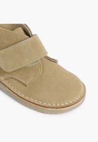 Beige semišová bota se suchým zipem, kulatou špičkou a prošívanými detaily podél podrážky pro větší odolnost.