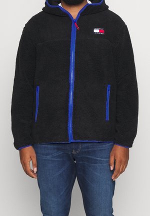 Mand iført en sort Sherpa lynlåstrøje med blå kant, lynlåslommer i siderne og Tommy Jeans-logo på brystet, kombineret med blå jeans.