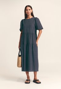 Abito in cotone blu navy con design a strati, maniche a sbuffo corte e sottili motivi verticali. La modella tiene una borsa intrecciata e indossa sandali neri.