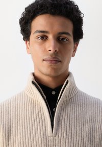 Suéter beige de canalé con un alto cuello, que presenta un cierre de media cremallera y botones oscuros contrastantes debajo. Textura suave y diseño ajustado.