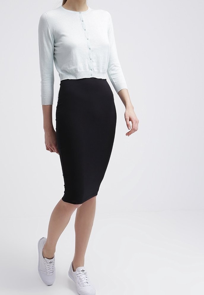 Femme portant un cardigan blanc court, une jupe crayon noire arrivant aux genoux et des baskets blanches, debout devant un fond clair uni.