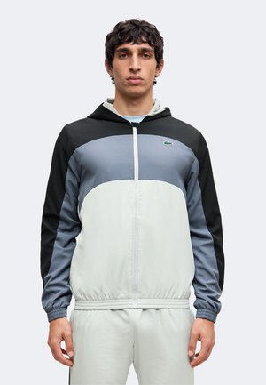 Jeune homme aux cheveux courts et bouclés portant une veste à capuche zippée noire, grise et blanche et un pantalon clair sur un fond uni.