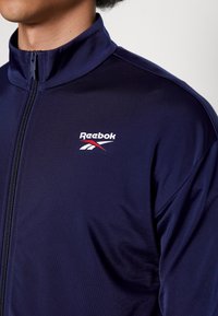 Námořnicky modrá bunda na zip z hladké látky s vysokým límcem. Na levé straně hrudi je logo Reebok v bílé barvě s červenými a modrými akcenty.