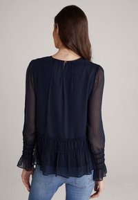 Dunkelblaue Bluse mit langen transparenten Ärmeln, gerafften Details an den Bündchen und am Saum sowie einem gerafften Ausschnitt mit einem kleinen Knopfverschluss auf der Rückseite.