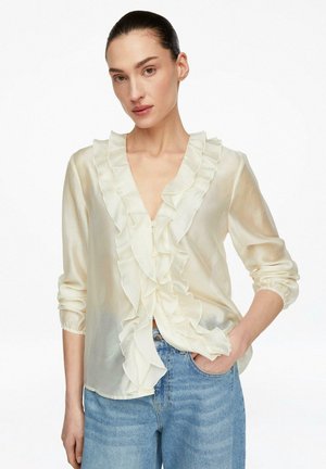 Kobieta ubrana w przejrzystą, kremową bluzkę z falbankami z przodu i długimi rękawami oraz jasnoniebieskie dżinsy, stojąca z jedną ręką w kieszeni.