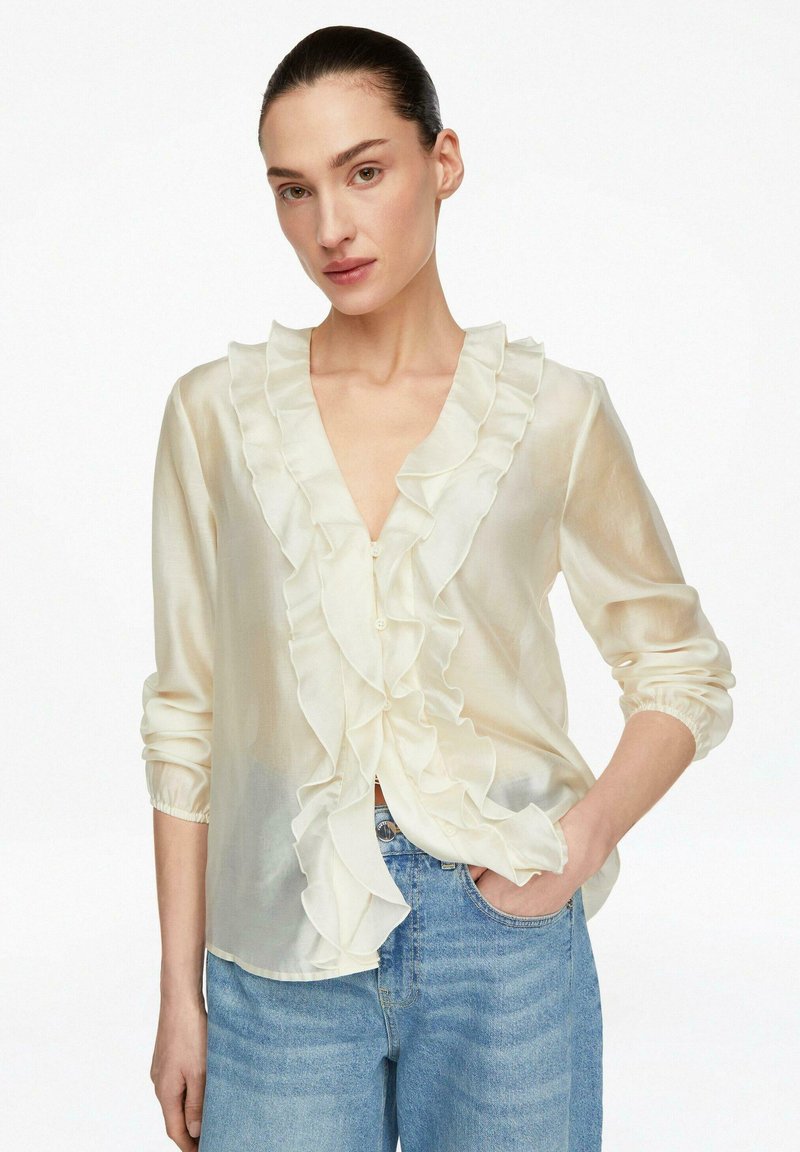 Vrouw die een doorschijnende crèmekleurige blouse met ruches aan de voorkant en lange mouwen draagt, gecombineerd met lichtblauwe spijkerbroek, staand met één hand in de zak.