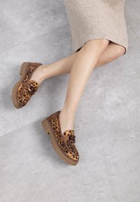 Zapatos de cuero con estampado de leopardo y acento de cuero marrón, suela gruesa, con un nudo decorativo en la parte frontal, sobre una superficie gris clara.