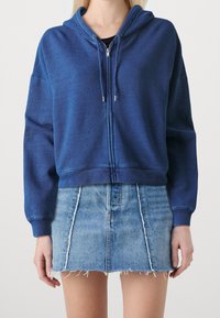 Sudadera azul con capucha y cremallera, de tejido texturizado y cordones, combinada con una minifalda de mezclilla deshilachada.