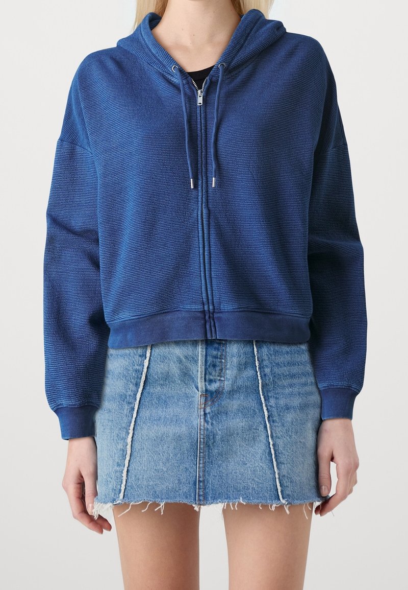 Sudadera azul con capucha y cremallera, de tejido texturizado y cordones, combinada con una minifalda de mezclilla deshilachada.