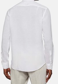 Camicia bianca a maniche lunghe realizzata in tessuto leggero. Presenta un colletto con bottoni, un inserto posteriore e spaccature laterali sull'orlo.