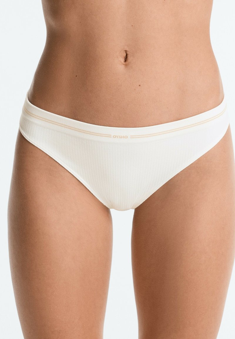 Personne portant un sous-vêtement blanc côtelé type bikini avec une bande horizontale subtile près de la taille, visible de la taille au haut des cuisses.