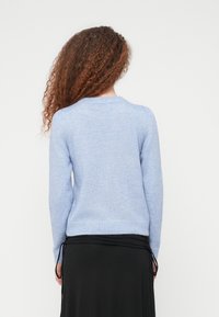 Άτομο με σγουρά μαλλιά που φοράει ανοιχτόγαλου πουλόβερ με μακριά μανίκια και μαύρη φούστα, γυρισμένο πλάτη σε απλό λευκό φόντο.