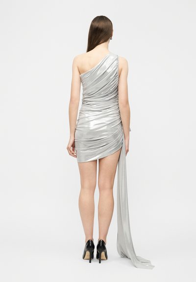Γυναίκα όρθια, που φοράει ένα ασημένιο μεταλλικό μίνι φόρεμα με έναν ώμο και με υφασμάτινο draping που εκτείνεται από την πλευρά, συνδυασμένο με μαύρα ψηλά τακούνια.
