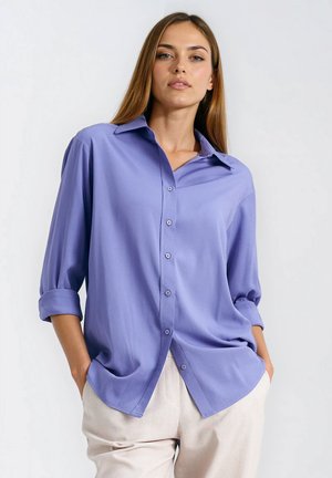 Lawendowa koszula na guziki z kołnierzykiem, rękawy podwinięte, o luźnym kroju. Wykonana z gładkiego materiału o subtelnym połysku, bez widocznych wzorów.