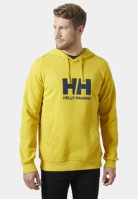 Sudadera con capucha amarilla, con un bolsillo frontal y el logo "HH" en azul marino. Cuenta con capucha con cordón y puños acanalados. Textura de tela suave.