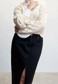 Pull en tricot cr ème épais avec motif en losange, superposé à une chemise blanche, associé à une jupe en jean noire avec une fente sur la cuisse.