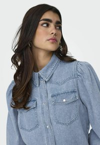 Camicia di jeans azzurro chiaro con colletto, chiusura a bottoni, maniche a sbuffo e due tasche sul petto con bottoni argentati. Texture liscia.