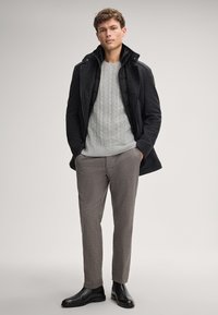 Herrenoutfit mit einem dunkelgrauen Mantel mit schwarzem Innenfutter, einem hellgrauen grob gestrickten Pullover und strukturierten grauen Hose. Schwarze Knöchelstiefel.