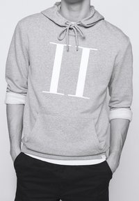 Sudadera con capucha gris con un gran gráfico blanco "II". Presenta un cierre con cordón, bolsillo frontal y un ribete blanco en contraste en las mangas.