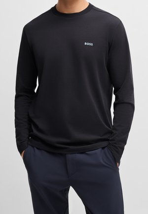 Mężczyzna ubrany w czarną koszulkę z długim rękawem i okrągłym dekoltem, z małym białym logo "BOSS" na piersi, ręce w kieszeniach, widoczne granatowe spodnie.