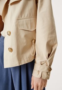 Beżowa kurtka z podwójnym zapięciem z przodu, dużą kieszenią na klatce piersiowej i mankietami na guziki. Posiada detal ze sznurkiem w okolicy dołu.