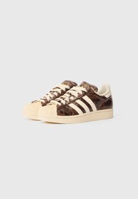 Sneakers castanhos com textura e padrão de réptil, apresentando três riscas brancas, uma sola bege e um design de bico em concha. Os atacadores creme completam o visual.