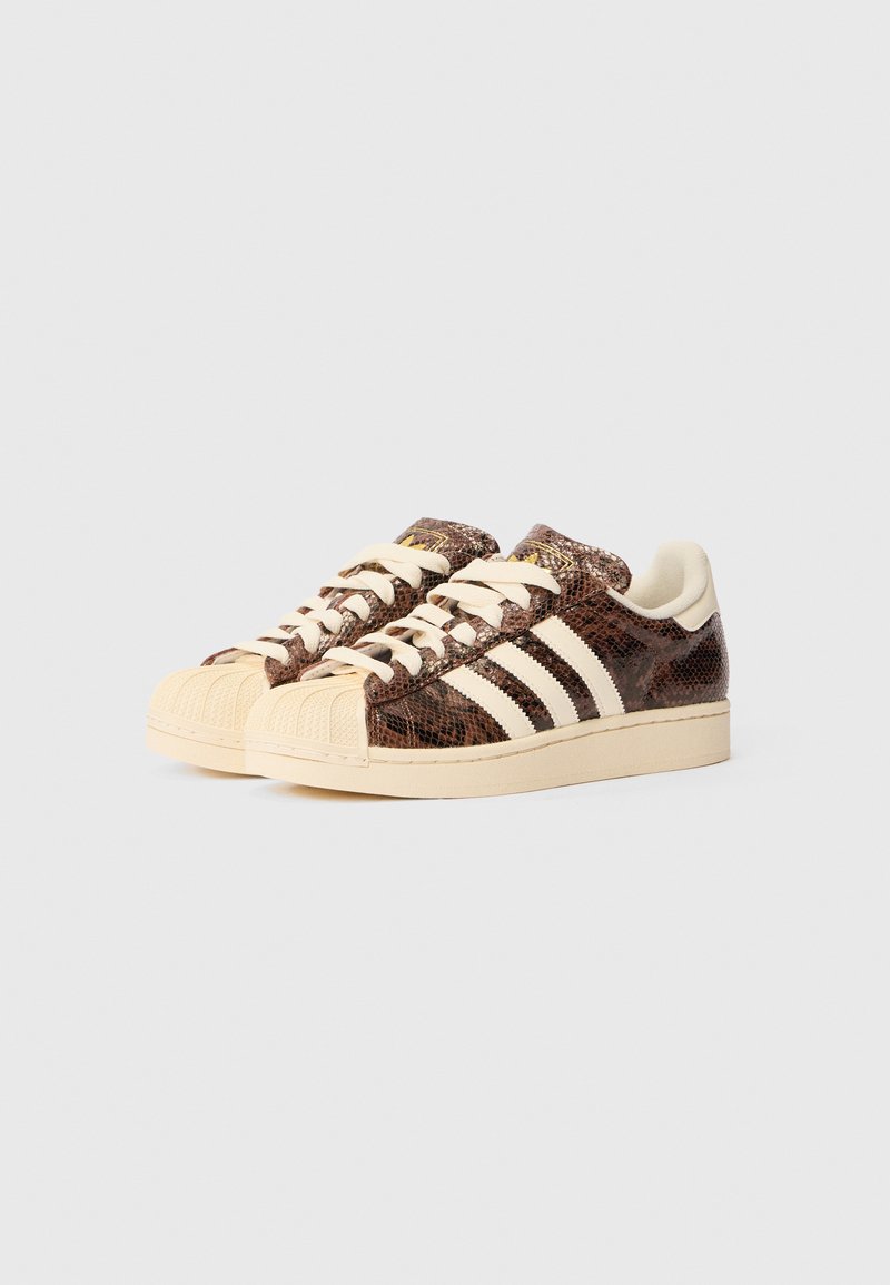 Baskets texturées marron avec un motif reptilien, dotées de trois rayures blanches, d'une semelle beige et d'un design à bout en coque. Des lacets crème complètent le look.