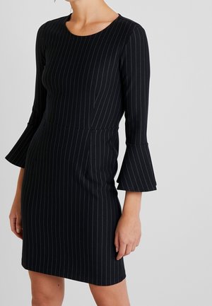 Femme portant une robe noire à fines rayures avec des manches évasées aux trois quarts, debout devant un fond gris clair uni.