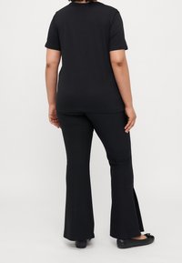 Person som bär en svart kortärmad skjorta, svarta utsvängda byxor med sidofickor och svarta ballerina skor, stående mot en enkel vit bakgrund.