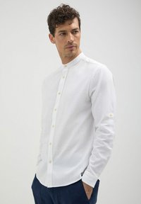 Camicia bianca a maniche lunghe con colletto alla coreana, presenta una chiusura frontale con bottoni, tessuto testurizzato e polsini a un bottone. Senza fantasie né accenti.