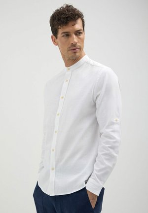 Camicia bianca a maniche lunghe con colletto alla coreana, presenta una chiusura frontale con bottoni, tessuto testurizzato e polsini a un bottone. Senza fantasie né accenti.