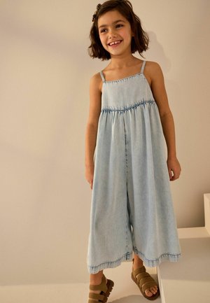 Junges Mädchen lächelt, trägt einen hellblauen Denim-Overall mit weitem Bein und braunen Sandalen, steht vor einem schlichten beigen Hintergrund.