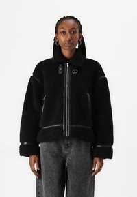 Zwarte fleece jas met leren accenten, ritsluiting en een hoge kraag. Heeft brede mouwen en ribbelmanchetten. Gedragen over grijze denim broeken.