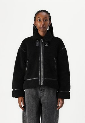 Chaqueta de forro polar negra con detalles de cuero, cierre de cremallera y cuello alto. Presenta mangas anchas y puños de canalé. Lleva puesta sobre pantalones de mezclilla gris.