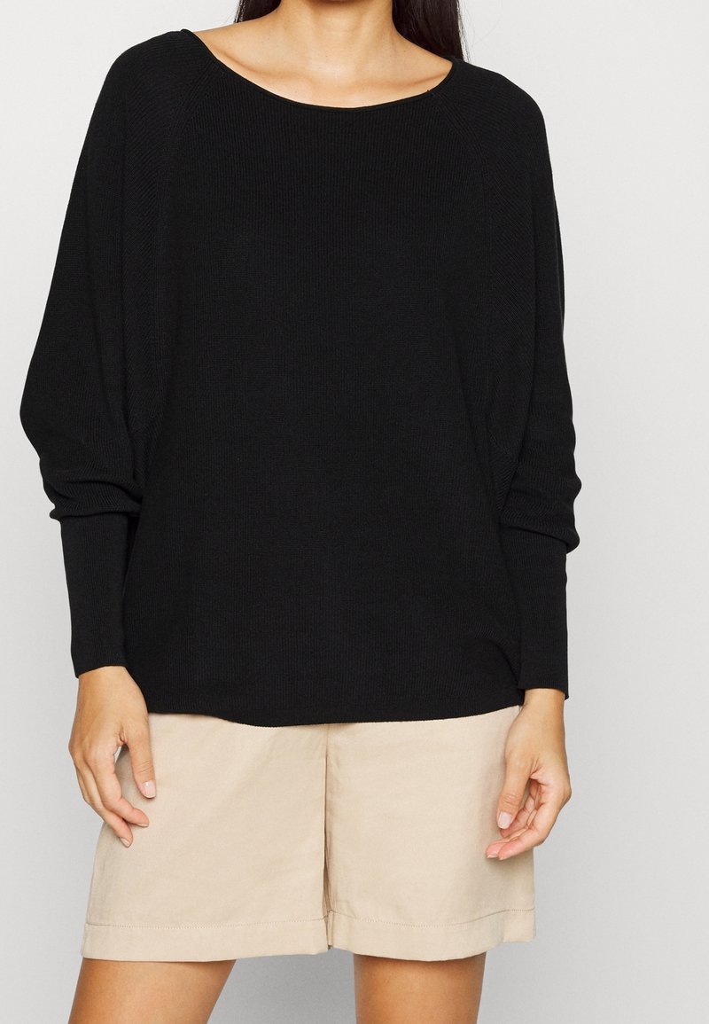 Schwarzer, oversized Strickpullover mit weitem U-Bootausschnitt und langen Ärmeln, kombiniert mit hellbeigen Shorts. Weiche Textur mit gerippten Details.