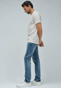 Hombre con una camisa blanca de manga corta, vaqueros azules y zapatillas blancas de pie de perfil con las manos entrelazadas detrás de la espalda, contra un fondo sencillo.