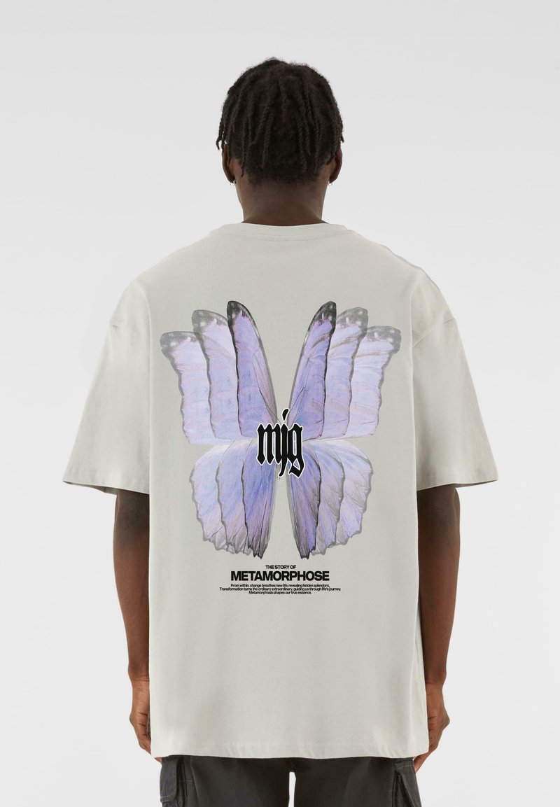 Lys grå oversized t-shirt med et stort lilla sommerfugl-grafik på ryggen, med sort tekst nedenfor og en glat bomuldsstruktur.