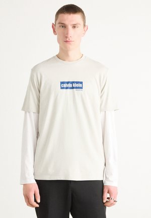 Lichtgrijze T-shirt met een blauw en wit "calvin klein" grafisch ontwerp. Draag over een witte longsleeve. Eenvoudig ontwerp, korte mouwen.