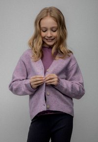 Młoda dziewczyna o blond włosach zapinająca lawendowy kardigan na fioletową bluzkę, stojąca na jednolitym szarym tle.