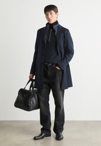 Bolsa de couro preta, casaco azul-marinho com botões, suéter azul-marinho com fecho, jeans escuros e botas de couro pretas. Texturas suaves e corte ajustado.