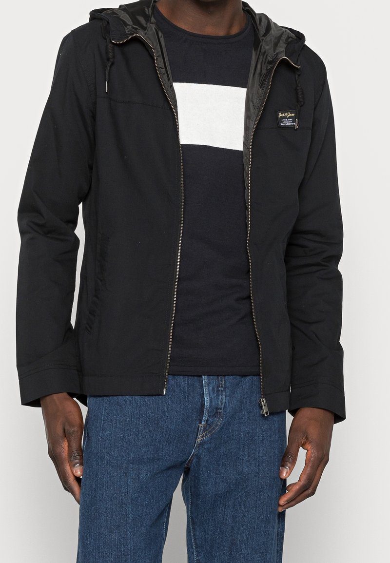 Giacca nera con cappuccio realizzata in tessuto leggero, con zip frontale, polsini elastici e una fodera interna liscia. Abbinata a una camicia blu navy.