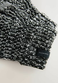 Pleteni črno-beli zimski klobuk z teksturiranim vzorcem in majhno črno etiketo, na kateri piše "ALLSAINTS". Mehki, topli materiali.