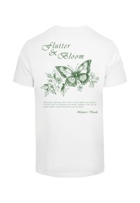 Hvid t-shirt med grønne sommerfugle- og blomsterdesigns på ryggen. Teksten siger "Flutter & Bloom" med yderligere dekorative elementer.