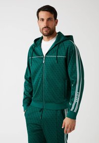 Sudadera verde con cremallera y un patrón texturizado, líneas blancas en los hombros y marca en el brazo izquierdo. Cuenta con cordones ajustables.