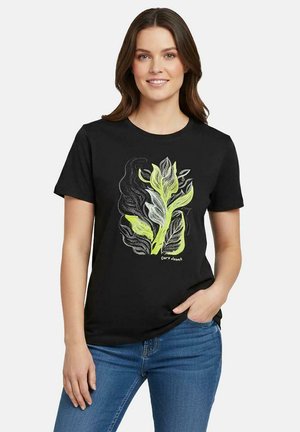 Vrouw draagt een zwart T-shirt met een abstract geel en grijs bladmotief, gecombineerd met een blauwe spijkerbroek, glimlachend met één hand in haar zak.